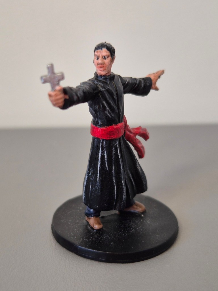 Figura di un sacerdote con un crocifisso, vestito di nero con una cintura rossa, su una base nera.