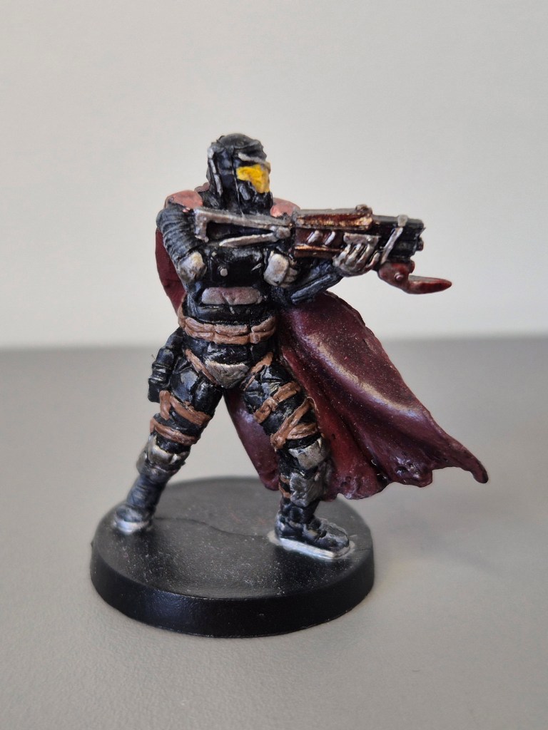 Miniatura di un guerriero con una maschera nera e occhiali gialli, vestito con un mantello rosso e armato di fucile, posizionato su una base nera.