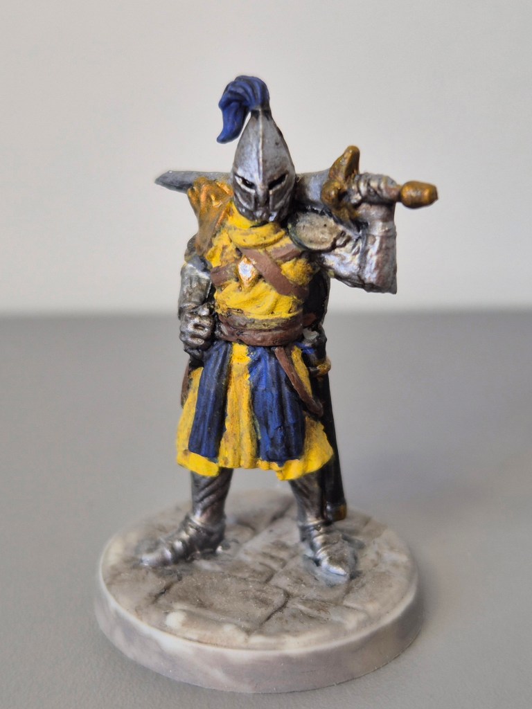 Una figura di un cavaliere con armatura, vestito di giallo e blu, impugnando una spada, posizionata su una base circolare.